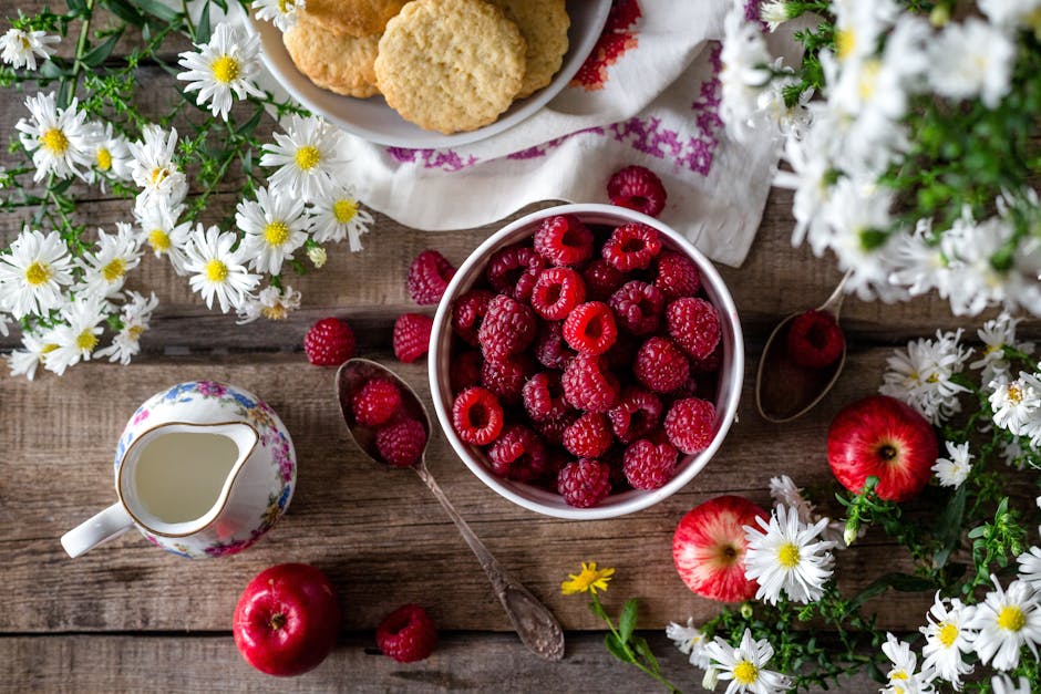 A rustic breakfast scene featuring a bowl of raspberries, apples, cookies, and white flowers on a wooden table