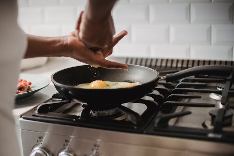 Close-up of frying eggs on a gas stove with hands seasoning the dish.
